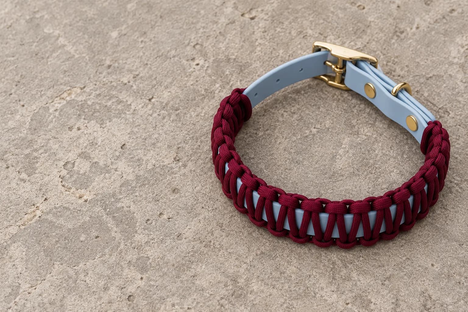 Halsband met bordeauxrood gevlochten koord over een lichtblauwe band, met messing gesp, op een natuurstenen ondergrond.